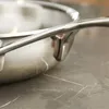 A close-up shot of the handle and rim of a stainless steel frying pan, resting on a marble countertop. Voir les images du produit
