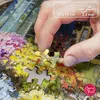 A hand is placing a puzzle piece into a larger puzzle depicting a colorful floral market scene. Podgląd zdjęcia produktu