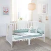 A white baby crib with mint green bedding and pillows, set in a nursery with artwork on the wall and a woven pendant light. Produktbild-Vorschau