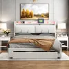 A bedroom scene featuring a white upholstered bed with a headboard that has built-in shelves and LED lighting. The bed is made with patterned bedding and a beige throw blanket. Two white nightstands with lamps are placed on either side of the bed. Produktbild-Vorschau
