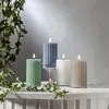 Four LED pillar candles of different colors and sizes are arranged on a white surface, with ivy vines draped around them. The background is a plain, light gray wall. Podgląd zdjęcia produktu