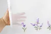 A hand is holding up a sheer white curtain with embroidered lavender flowers along the bottom edge. Produktbild-Vorschau