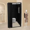 A black freestanding changing room with a stool inside, set against a light beige wall. A mannequin dressed in a beige vest and black top stands to the right. Produktbild-Vorschau