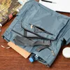 A close-up view of a gray handbag on a wooden surface, with a magazine, a notebook, a pen, a water bottle, and a small white container visible. Produktbild-Vorschau