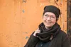 A woman wearing a knit hat and scarf stands in front of an orange, textured wall. Produktbild-Vorschau