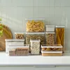 A collection of clear plastic food storage containers filled with various dry goods, arranged on a white countertop against a tiled backsplash. Produktbild-Vorschau