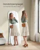 A woman is posing in front of a full-length mirror, checking her outfit. The mirror is tall and oval-shaped with a black frame, and it's standing on a black metal stand. The woman is wearing a light-colored skirt and a green shirt, and she has a brown purs Produktbild-Vorschau