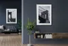 A living room with two framed pictures of a bridge on a dark blue wall, a sofa, a coffee table, a plant, and a wooden shelf with books and decorative items. Produktbild-Vorschau
