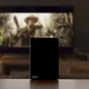 A black Vayox device is placed on a wooden surface in front of a blurred television screen displaying a scene from a movie. Podgląd zdjęcia produktu