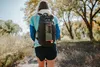 A person wearing a backpack with a water bottle attached is walking on a trail. Produktbild-Vorschau