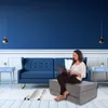 A woman sitting on a gray folding mattress and using a laptop in a living room with a blue sofa and blue walls. Náhled obrázku produktu