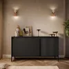 A modern living room with a black sideboard, decorative items, and wall sconces. Produktbild-Vorschau
