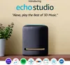 An Amazon Echo Studio speaker is shown on a wooden shelf next to a potted plant. The background is a plain, light beige color. Several service logos are displayed at the bottom of the image. Anteprima dell’immagine prodotto
