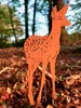 A metal deer figurine stands in a forest floor covered with fallen autumn leaves. Produktbild-Vorschau