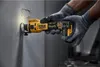 A person wearing gloves is using a DeWALT cordless drywall router to cut a hole in a wall. Podgląd zdjęcia produktu