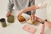 Two people are shown interacting with lunchboxes and bottles on a white table. Produktbild-Vorschau