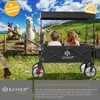 Two children are sitting in a KESSER bollerwagen (wagon) in a grassy field with alpacas in the background. Produktbild-Vorschau