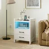A white nightstand with two drawers and an open shelf illuminated by blue LEDs, placed next to a sofa in a living room. Produktbild-Vorschau