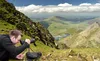 A man is using a spotting scope on a grassy mountainside overlooking a valley with lakes. Produktbild-Vorschau