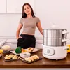 A woman stands in a kitchen next to a steaming appliance, with prepared food on the counter. Produktbild-Vorschau