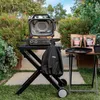 A Ninja Woodfire grill stand is set up outdoors in a garden setting, with the grill on top and a cover hanging from it. There are also bags of seasoning and a tray on a side table. Náhľad obrázku produktu