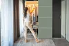 A woman is standing in front of an open closet, choosing clothes. The closet is well-lit and organized with shelves and hangers. Curtains are visible in the background. Produktbild-Vorschau