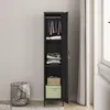 A tall, narrow black metal locker with its door open, revealing shelves with folded clothes, a basket, and a box. A wire basket with a blanket is to the left. The locker is placed in a room with a light-colored wall and a window with curtains on the right. Produktbild-Vorschau