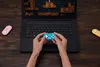 A person is playing a video game on a laptop with a small turquoise gamepad. Two other gamepads, pink and yellow, are on the table. Náhľad obrázku produktu
