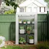 A white greenhouse with three shelves filled with plants, placed in a backyard next to a green fence and a house. Produktbild-Vorschau