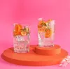 Two glasses of clear liquid with ice, orange slices, and rosemary sprigs are placed on an orange circular platform against a pink background. Anteprima dell’immagine prodotto