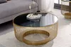 A round coffee table with a gold metal frame and a black glass top is placed on a rug in a living room setting. Produktbild-Vorschau