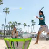 A man and a woman playing a paddle game on a beach with palm trees and buildings in the background. Produktbild-Vorschau