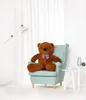 A large brown teddy bear sits in a light blue armchair in a brightly lit room with white curtains. Podgląd zdjęcia produktu