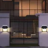 Two solar-powered wall lights are mounted on a white wall next to a dark metal gate. The lights are illuminated, casting a warm glow. Produktbild-Vorschau