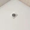 A close-up view of a white shower tray with a square stainless steel drain cover. Produktbild-Vorschau