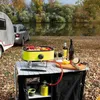 A campsite scene with a grill and food set up by a lake. Podgląd zdjęcia produktu