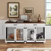 Two dogs are inside a white and brown dog crate furniture piece, which is placed on a wooden floor in front of a wall with artwork and decorative items. Produktbild-Vorschau