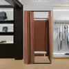 A freestanding changing room with brown curtains and a bench inside, set up in a retail store. Clothes are hanging on a rack to the right, and bags and shoes are displayed on shelves to the left. Voir les images du produit