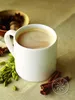 A white mug of chai tea is surrounded by spices like cardamom pods and cinnamon sticks, with a small bowl of star anise and nutmeg in the background. Produktbild-Vorschau
