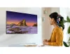 A woman is sitting at a desk, holding a mug and looking at a large TV screen mounted on the wall. The TV displays a scenic landscape image. Produktbild-Vorschau