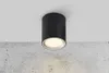 A black cylinder-shaped LED ceiling light is mounted on a white ceiling above a textured concrete wall. Produktbild-Vorschau