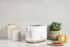 A white toaster with wood accents sits on a marble countertop next to two canisters and a potted plant. A slice of toast and a mug of tea are in the foreground. Náhled obrázku produktu