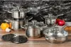 A set of stainless steel cookware arranged on a wooden surface with a dark marble-patterned background. Some vegetables (garlic, onion, tomato) are placed near the cookware. Voir les images du produit
