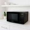 A black microwave oven is shown on a white countertop, with a small potted plant to its left and stacked white bowls and plates on a shelf above. Anteprima dell’immagine prodotto