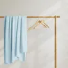 A light blue blanket is draped over a wooden hanger rack, with two empty wooden hangers also on the rack. The background is a plain wall with soft shadows cast by light coming from the side. Podgląd zdjęcia produktu