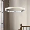 A modern circular pendant light fixture hangs in a well-lit interior space, possibly a hallway or staircase, with curtains and a wooden banister visible in the background. Produktbild-Vorschau