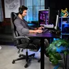 A man is playing a video game while sitting in a gaming chair in a room with a computer and neon lights. Produktbild-Vorschau
