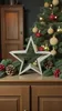 A wooden star decoration is placed on a wooden cabinet, with a Christmas tree and pine branches in the background. Produktbild-Vorschau