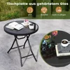 A round black folding side table with a glass top, shown outdoors on a patio. The table has books, a small cake, and a bowl of berries on it. A close-up inset shows flowers in a vase and a cup of coffee on a similar table. Produktbild-Vorschau