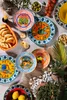 A collection of colorful decorative plates with various motifs, arranged on a textured surface with food items and greenery. Produktbild-Vorschau